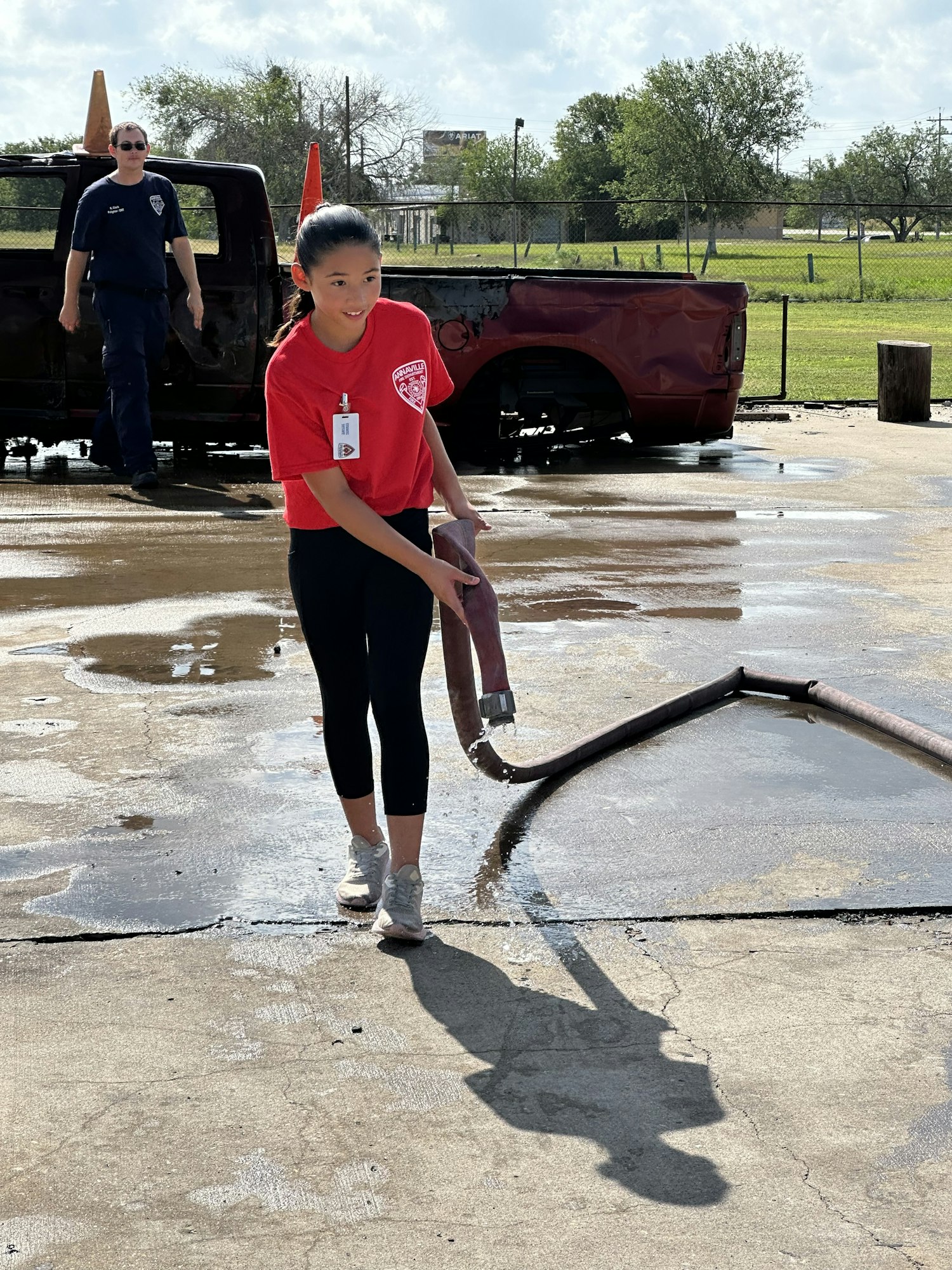 Person in a red shirt holding a hose on a wet surface, with a damaged vehicle and another person in the background.