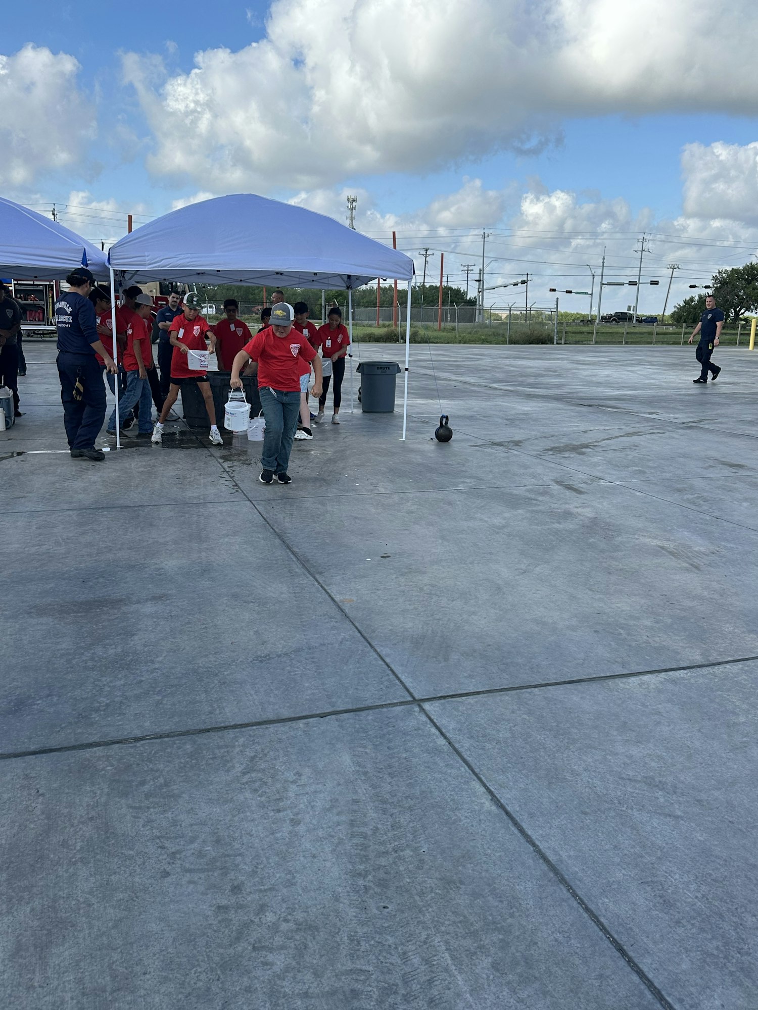 Group in red shirts assembling under a white canopy on a concrete area, with blue skies and clouds above.
