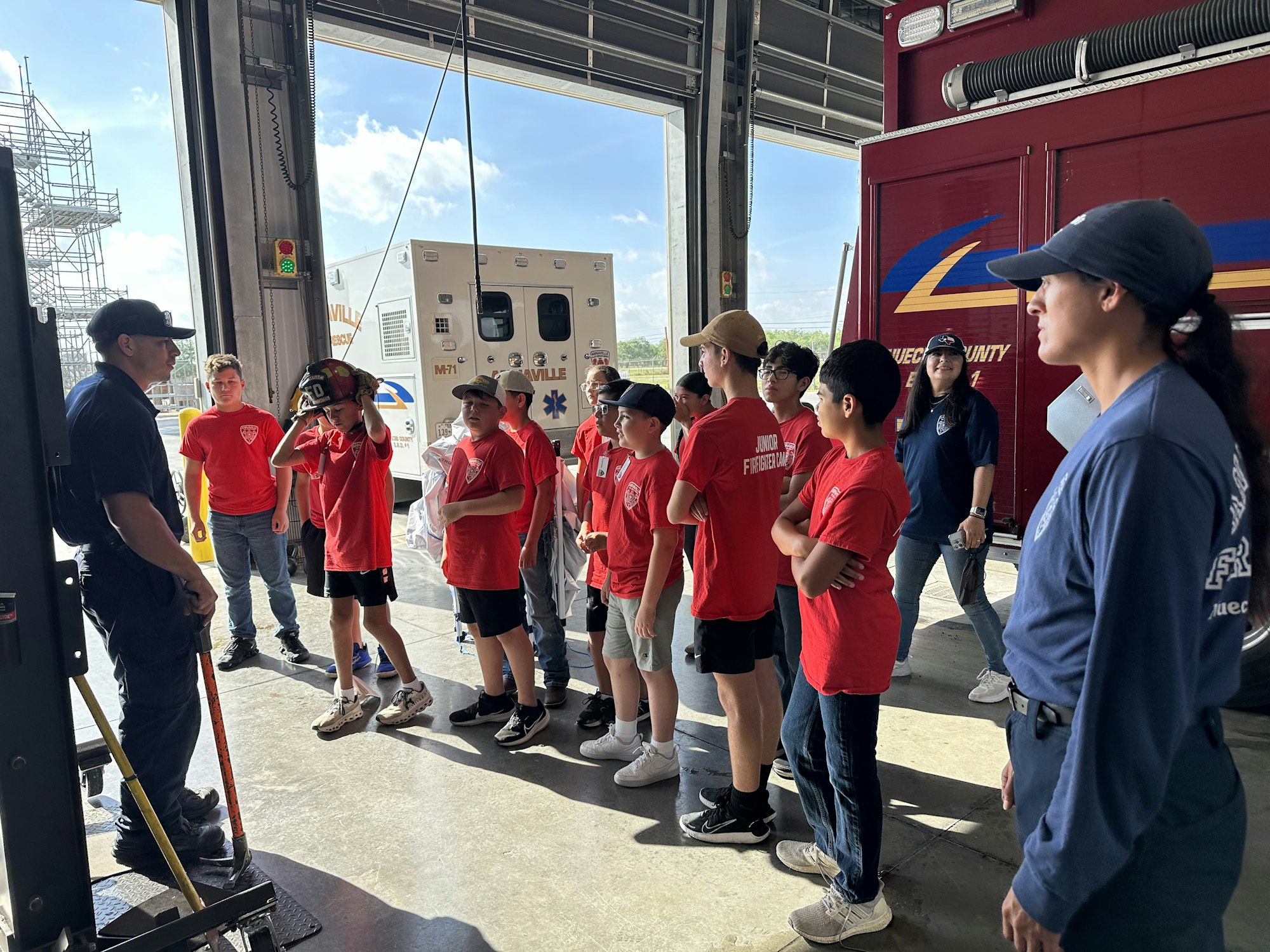 A firefighter is talking to a group of kids wearing red "Junior Firefighter Camp" shirts in a fire station with emergency vehicles.