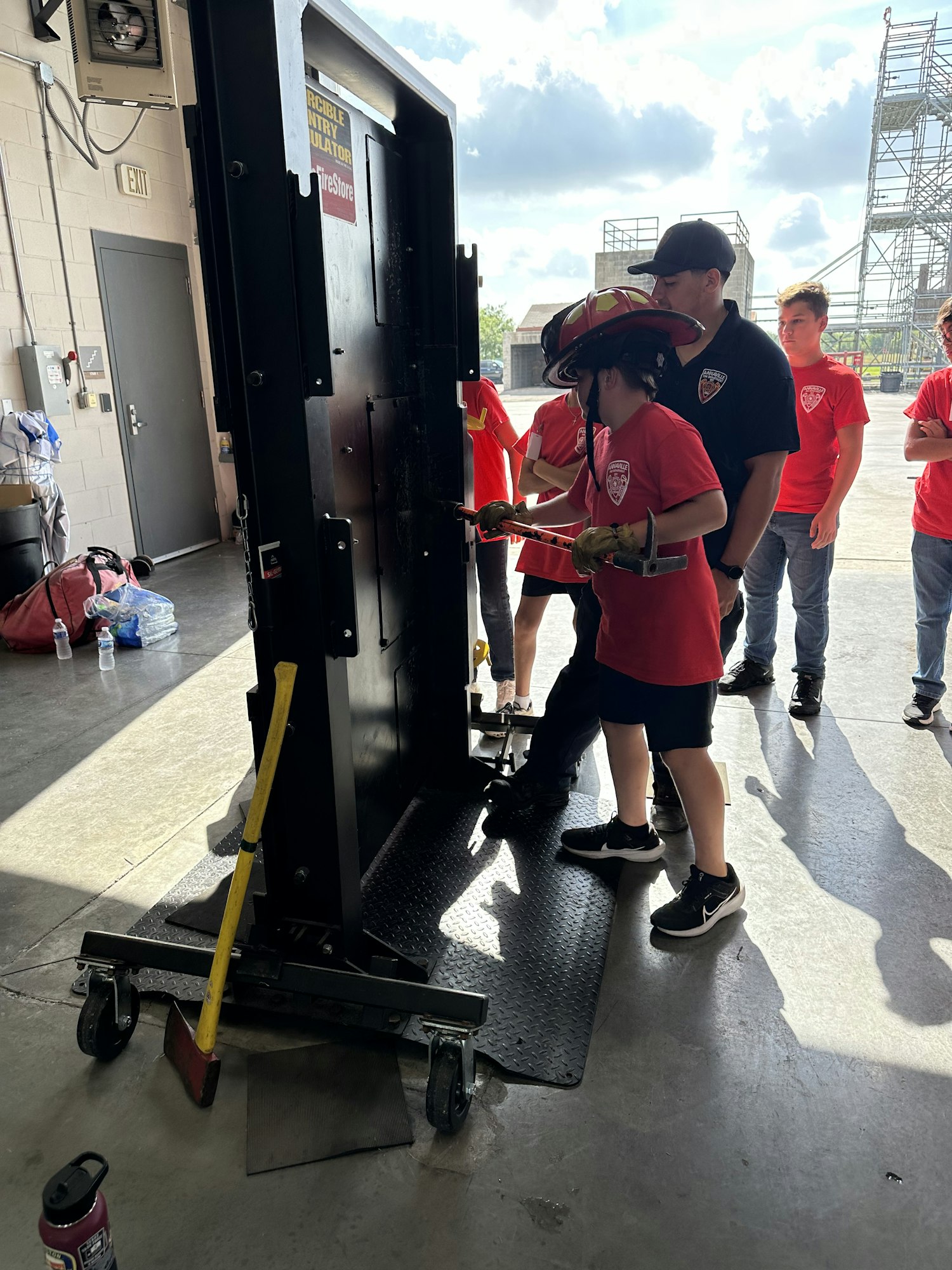 A person in a firefighter helmet practices using a tool on a training door, supervised by others in red shirts.