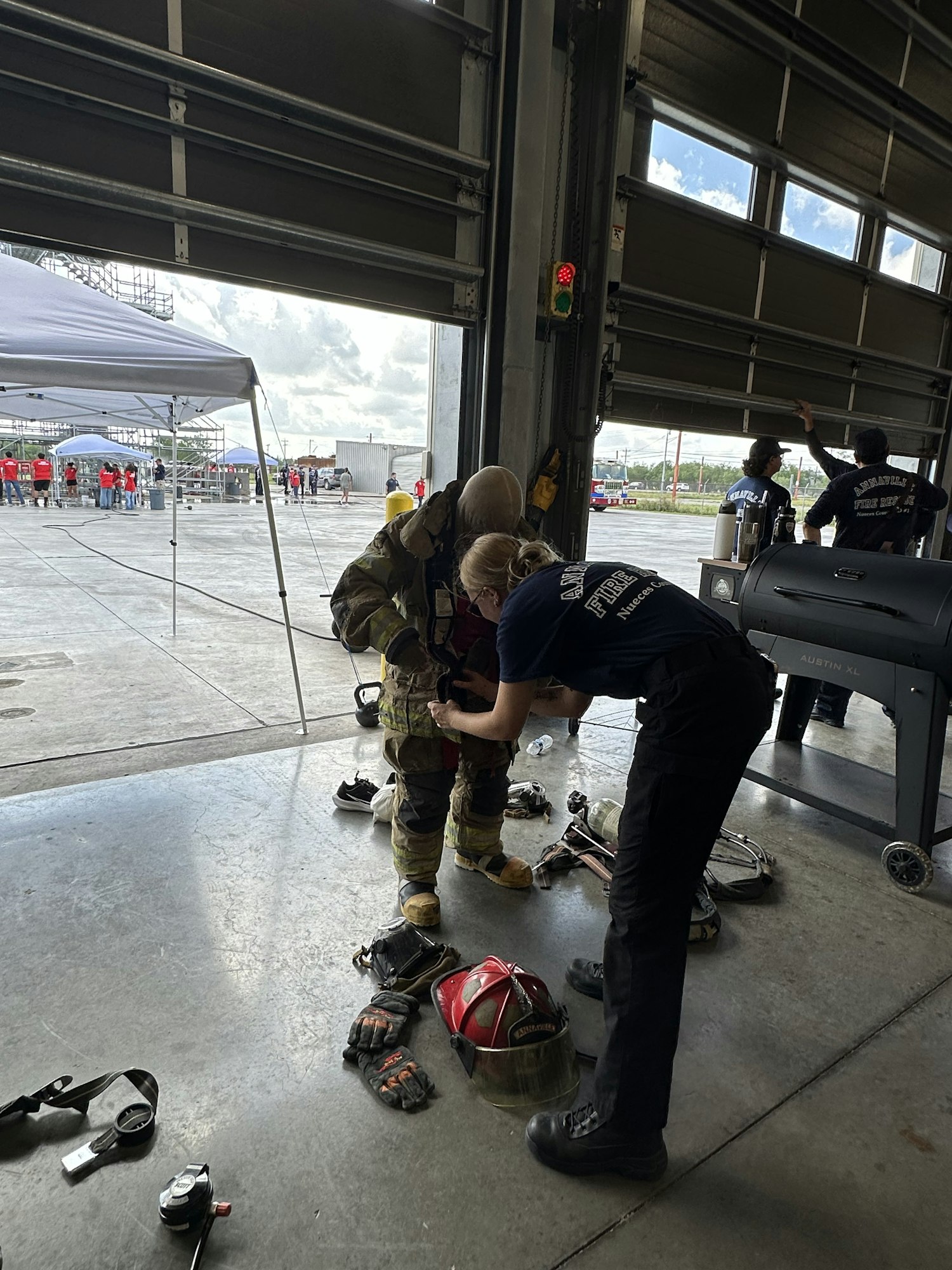 A firefighter is being assisted in a garage with gear on the floor. Equipment and people are visible outside near tents.