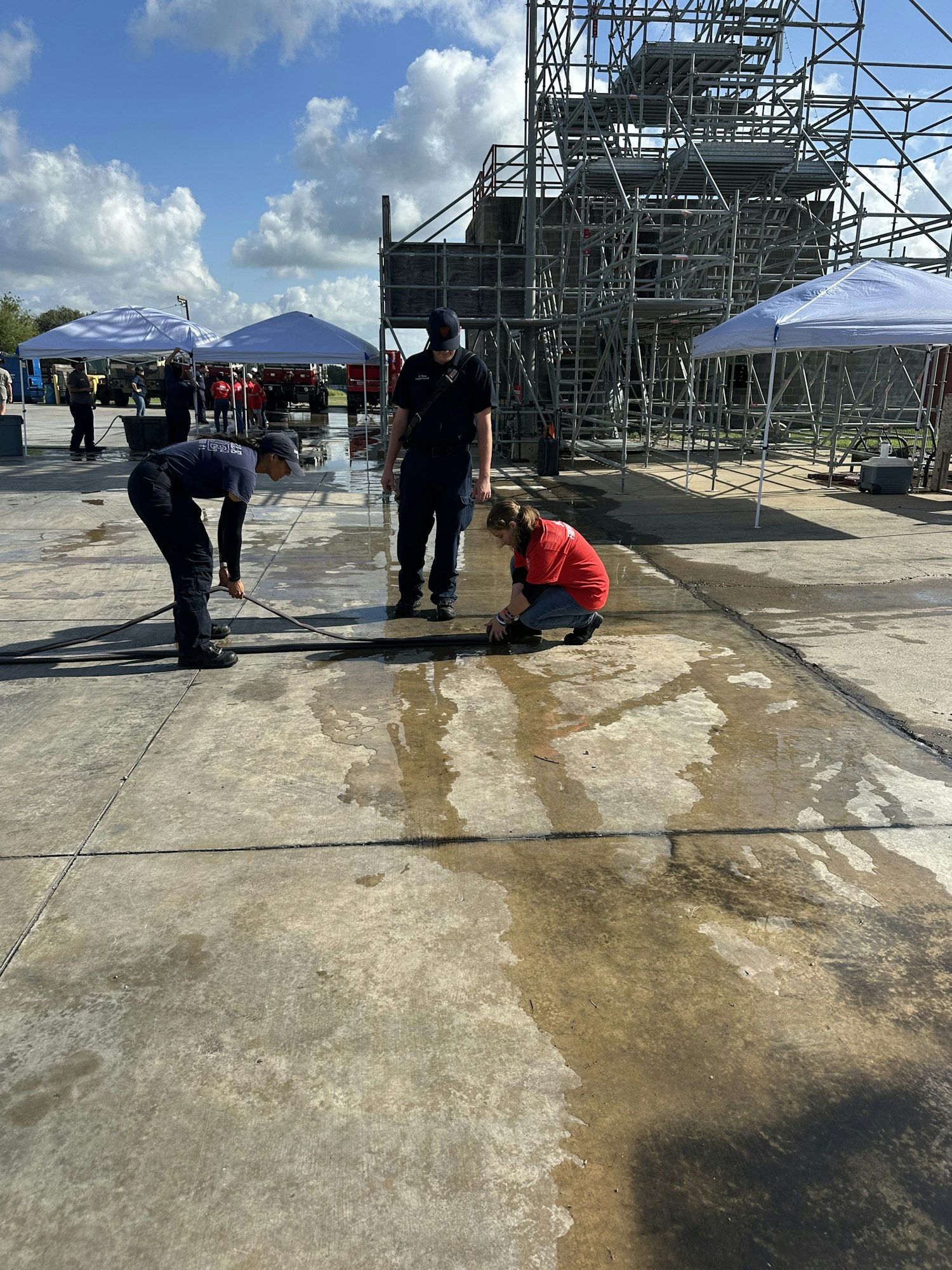 People cleaning a concrete surface with water hoses near a metal scaffolding structure and tents.