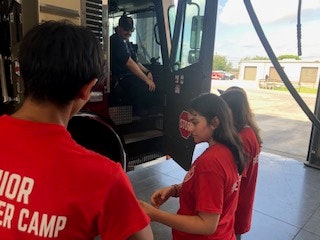 A group of people in red "Junior Firefighter Camp" shirts near a fire truck, with one person sitting inside the truck.