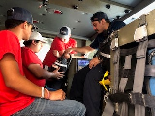 People sitting inside a vehicle; one in a uniform demonstrating equipment to three others in red shirts.