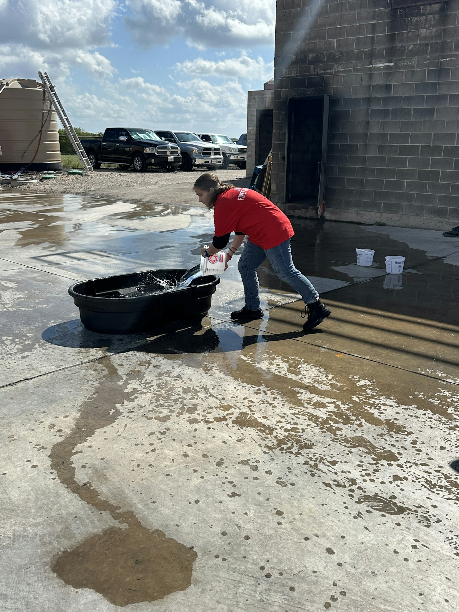 A person in a red shirt washes a bucket outdoors near a building, with scattered water on the concrete.