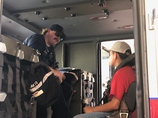 Two people inside a vehicle: one wearing a cap and sunglasses, the other in a red shirt and hat, sitting and talking.