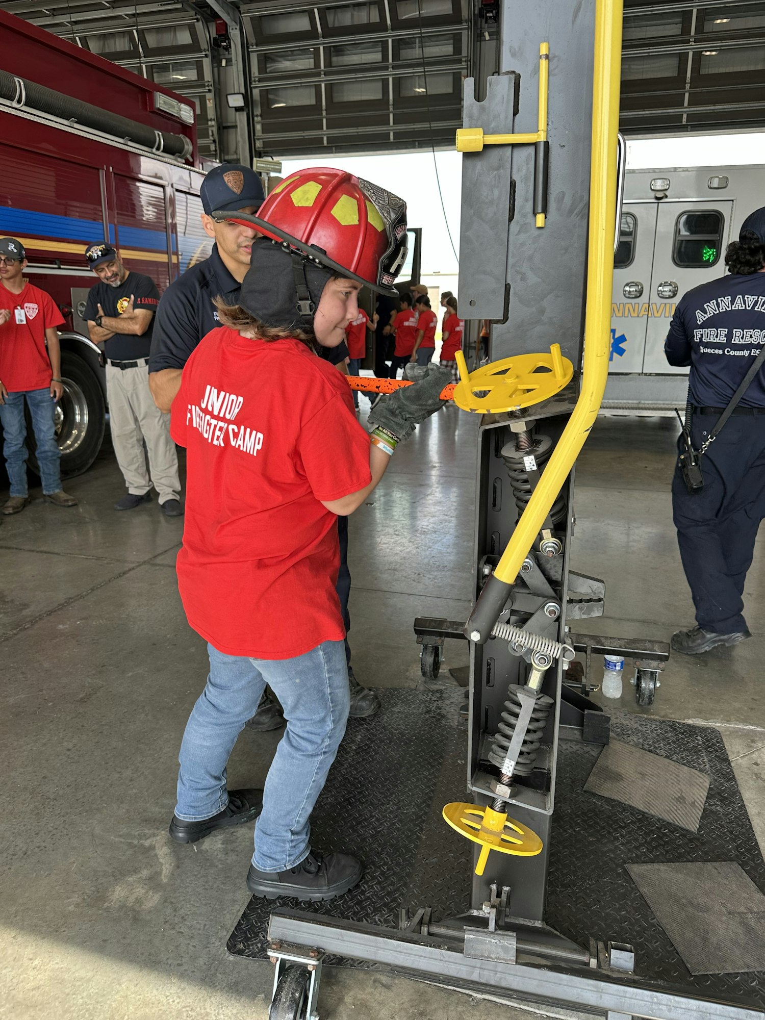 A person in a red helmet and shirt participates in an activity involving a mechanical apparatus, with fire personnel observing.