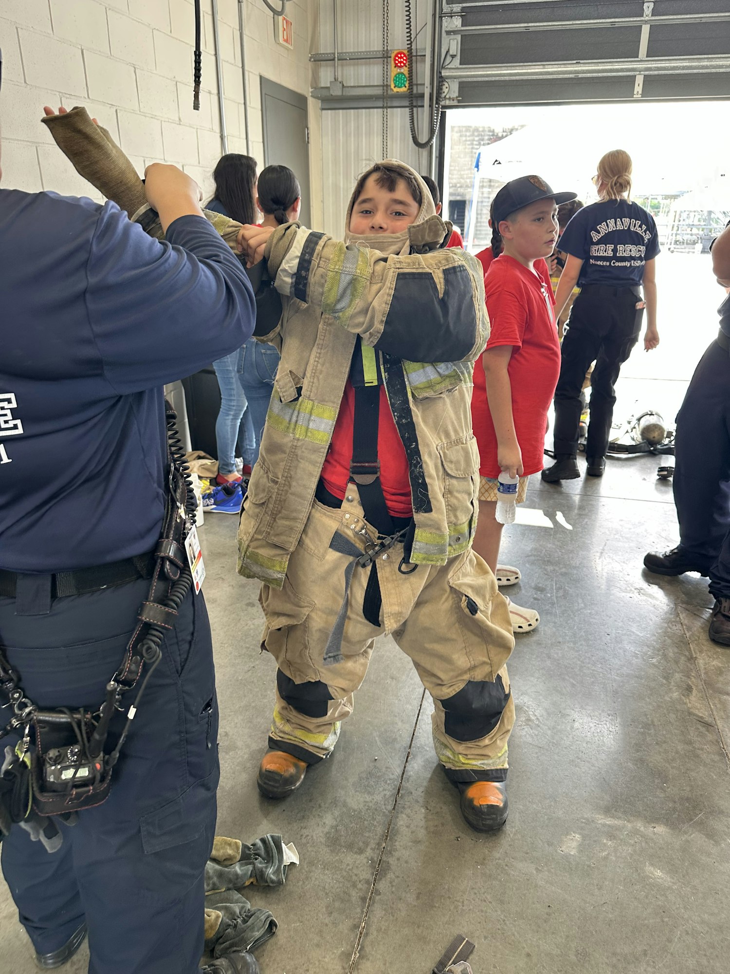A child in firefighter gear being assisted by an adult, surrounded by others in a garage setting.