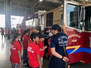 Children wearing red shirts are attending a fire station tour, guided by firefighters near a fire truck.