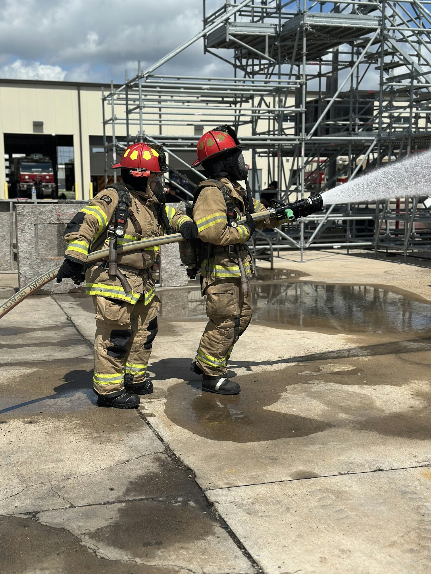 Two firefighters in full gear holding a hose, spraying water, with scaffolding and a building in the background.