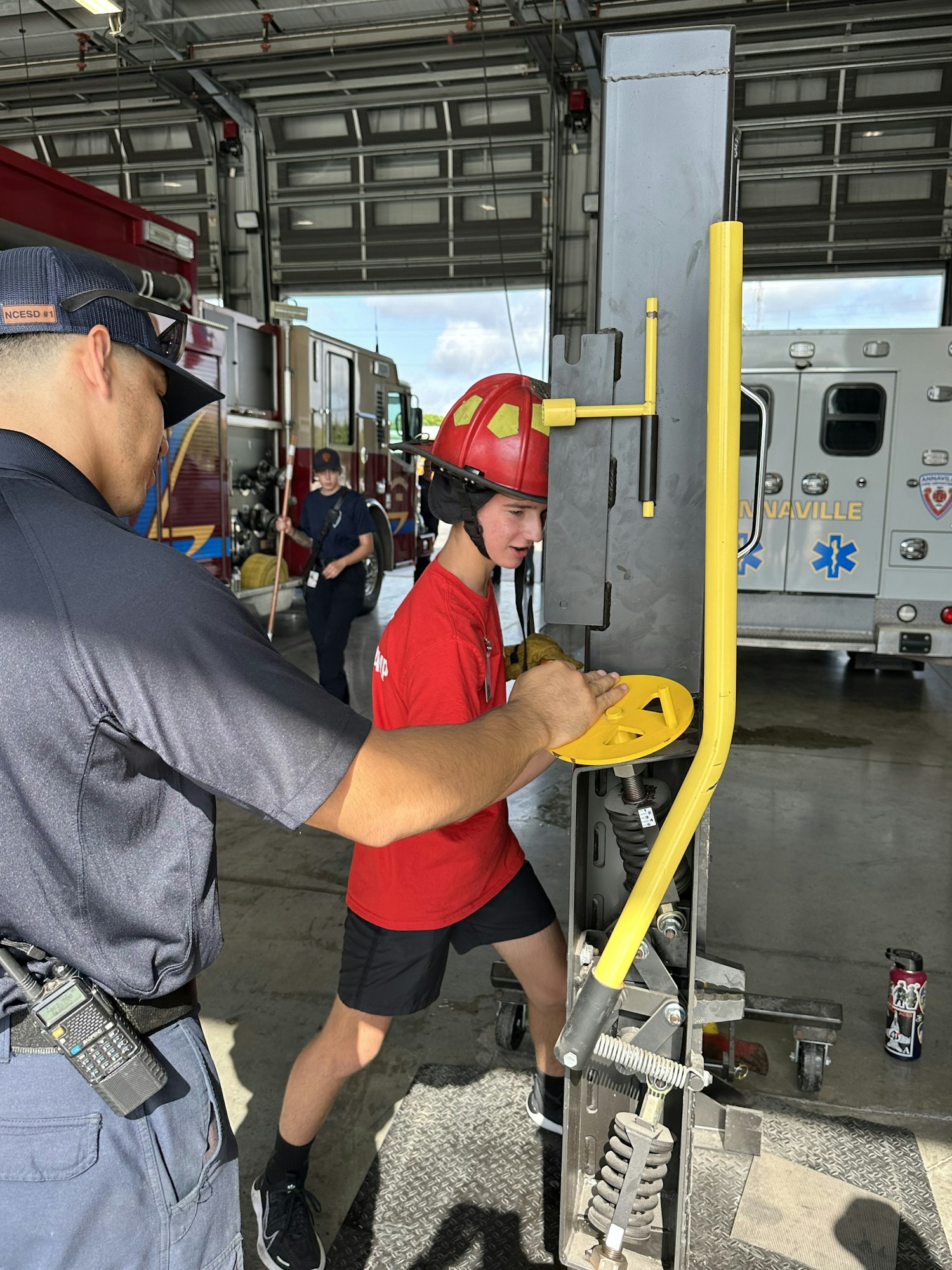 A person in a helmet uses a firefighting tool; another person watches in a fire station. Fire truck and ambulance in background.