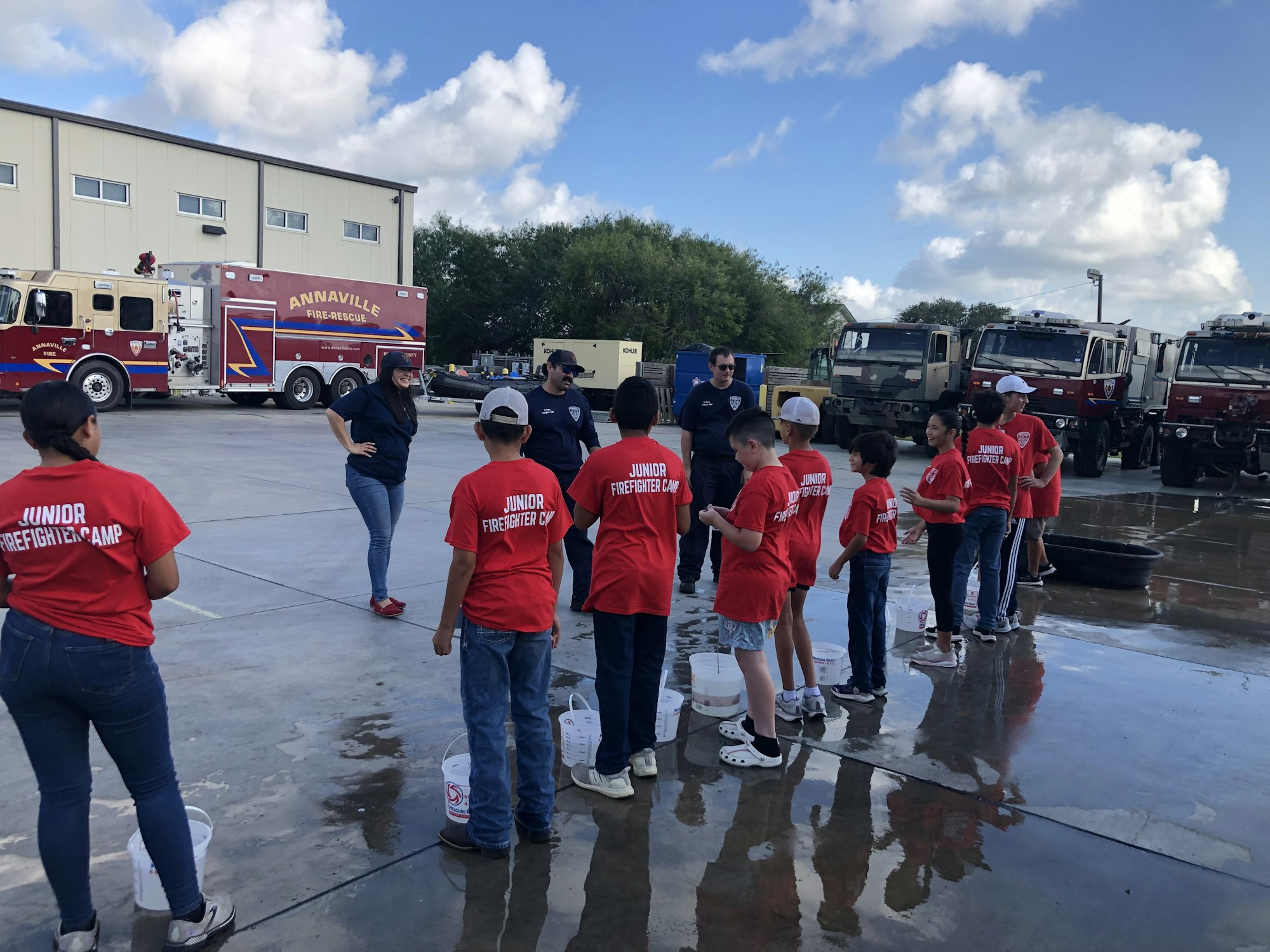 Children in red "Junior Firefighter Camp" shirts lined up, with fire trucks in the background, participating in an outdoor activity.