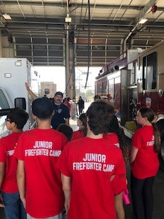 A group of people wearing red "Junior Firefighter Camp" shirts in a fire station, guided by someone in uniform.