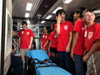 Group of teens in red shirts inside an ambulance, standing near a stretcher.