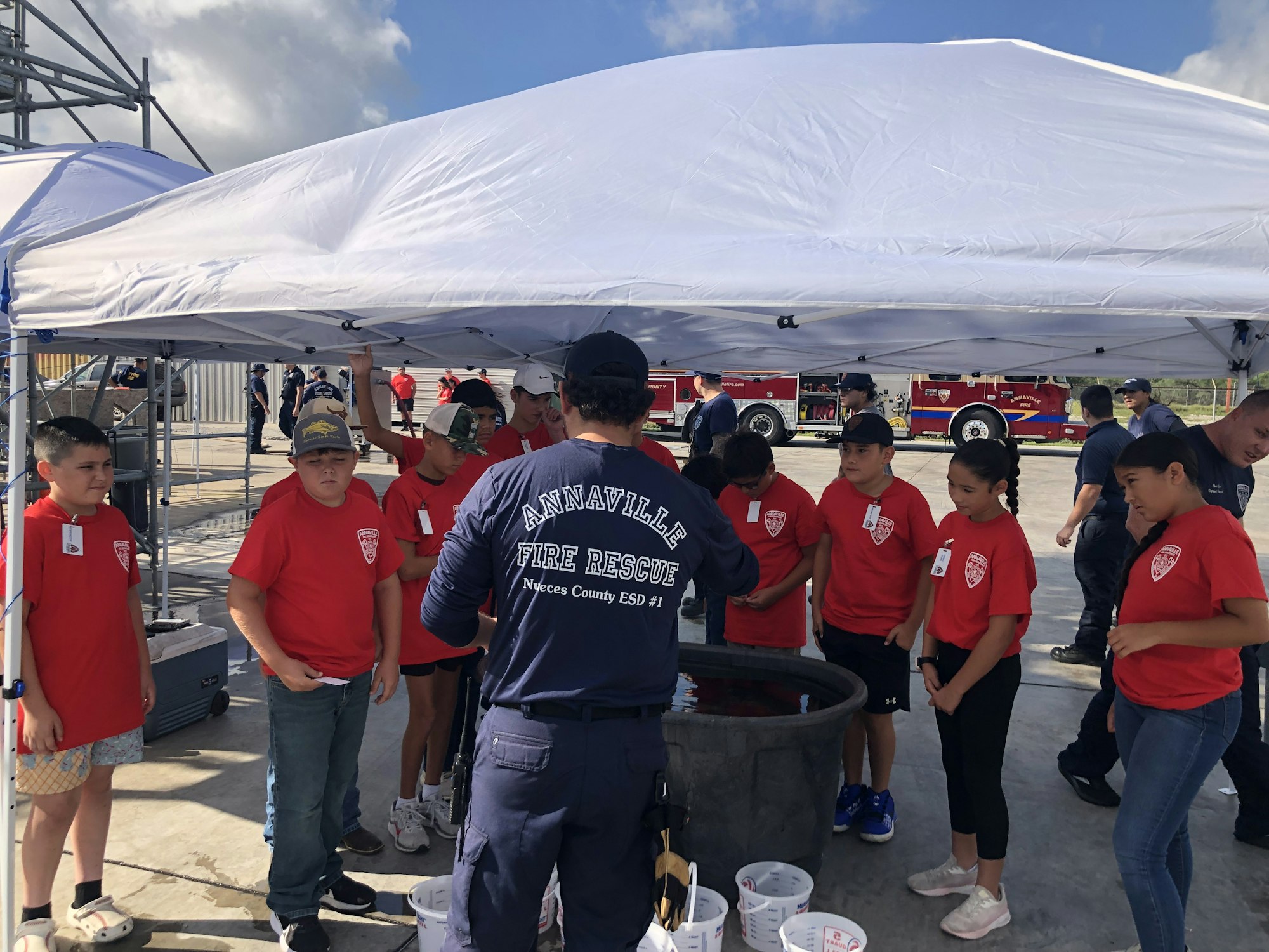 A group of children in red shirts gather under a canopy with a firefighter. Fire trucks are visible in the background.