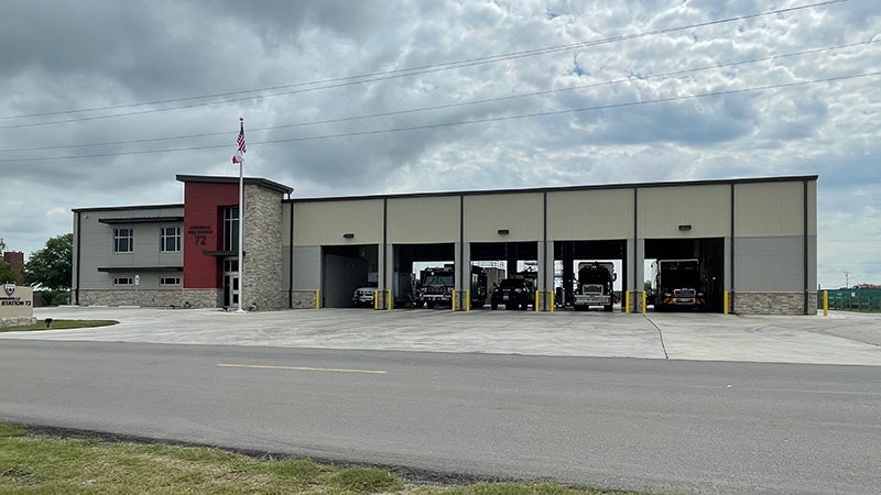 A modern fire station with several garage bays open, housing fire trucks, and an American flag pole nearby.