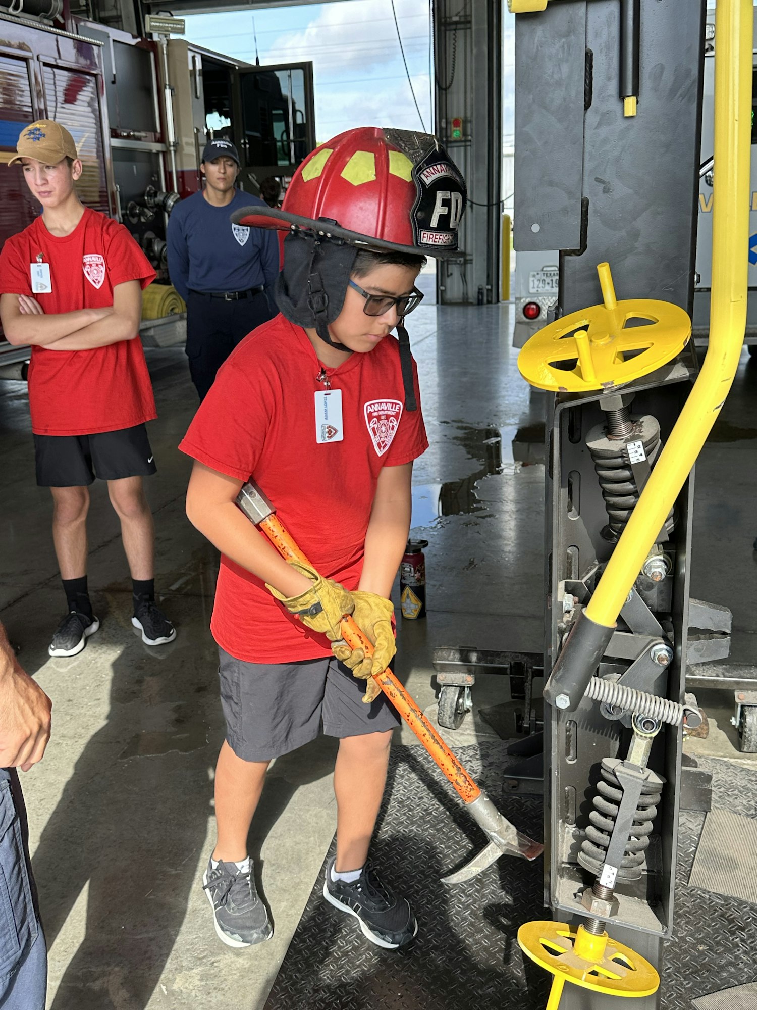 A person in firefighter gear holds a tool inside a station; others watch in the background.