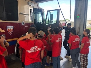 A group of kids wearing "Junior Firefighter Camp" shirts are gathered around a fire truck inside a fire station.
