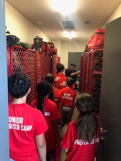 A group of people wearing red shirts in a locker room with red lockers, labeled "Junior Firefighter Camp."