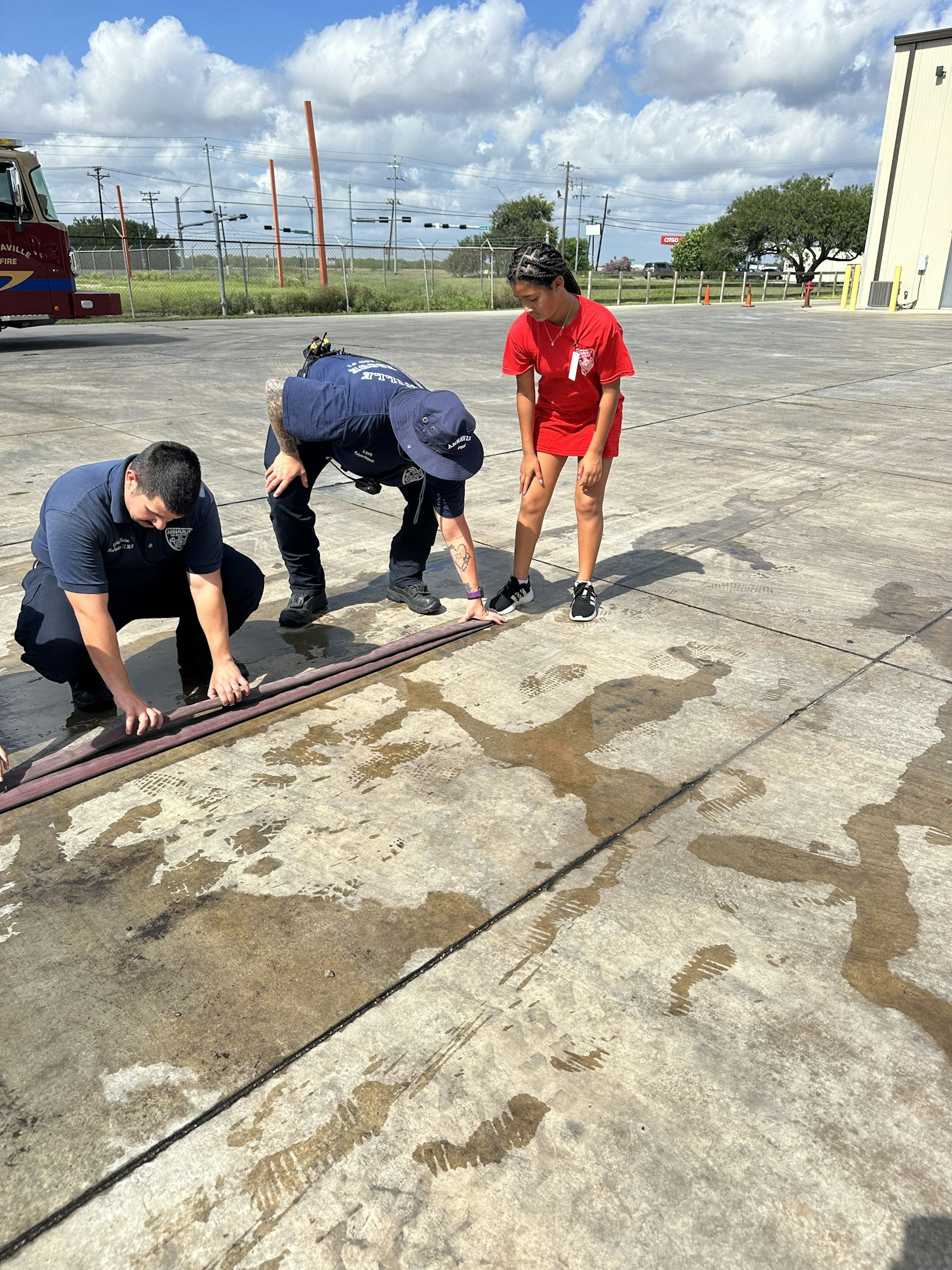Four people examining a hose on a wet concrete surface outdoors, with a fire truck in the background.