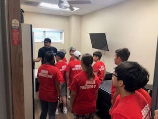 A group of kids in red shirts that say "Junior Firefighter Camp" listen to an adult in a room.