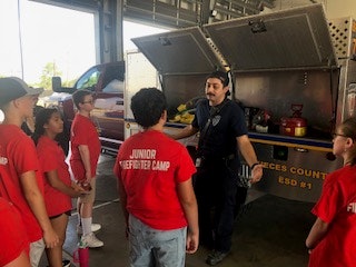 A firefighter talks to a group of children in red "Junior Firefighter Camp" shirts near an open fire truck compartment.