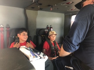 Two kids wearing seatbelts inside a vehicle, being talked to by an adult in uniform.