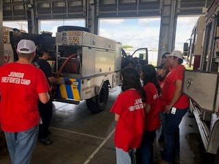 A group of people in red "Junior Firefighter Camp" shirts watch a small fire truck in a garage.