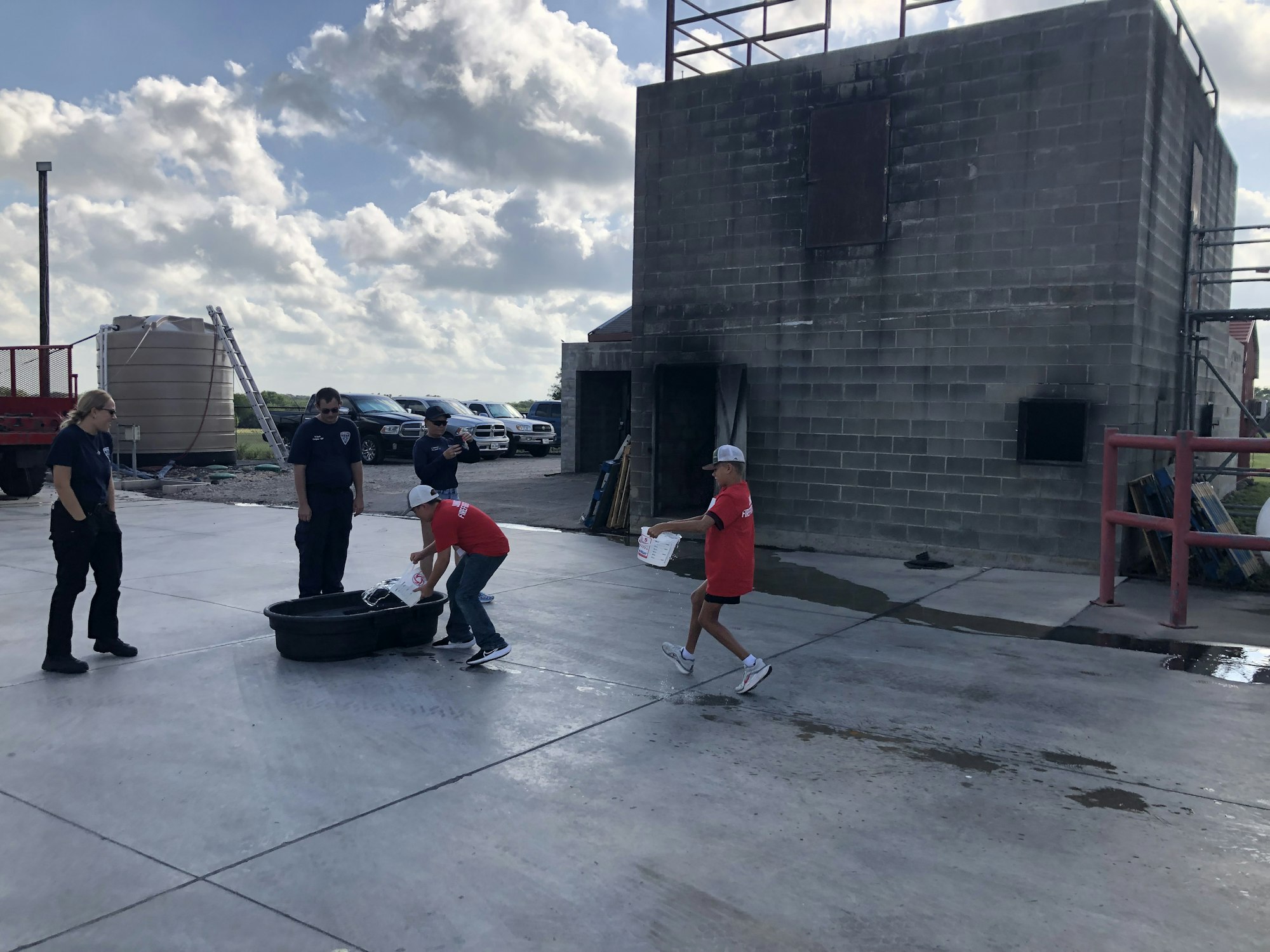 People in an outdoor area near a concrete building, with one person carrying a bucket. Several parked vehicles in the background.