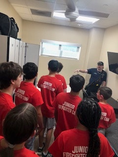 A group of people in red "Junior Firefighter" shirts listens to someone in a uniform in a room with lockers.