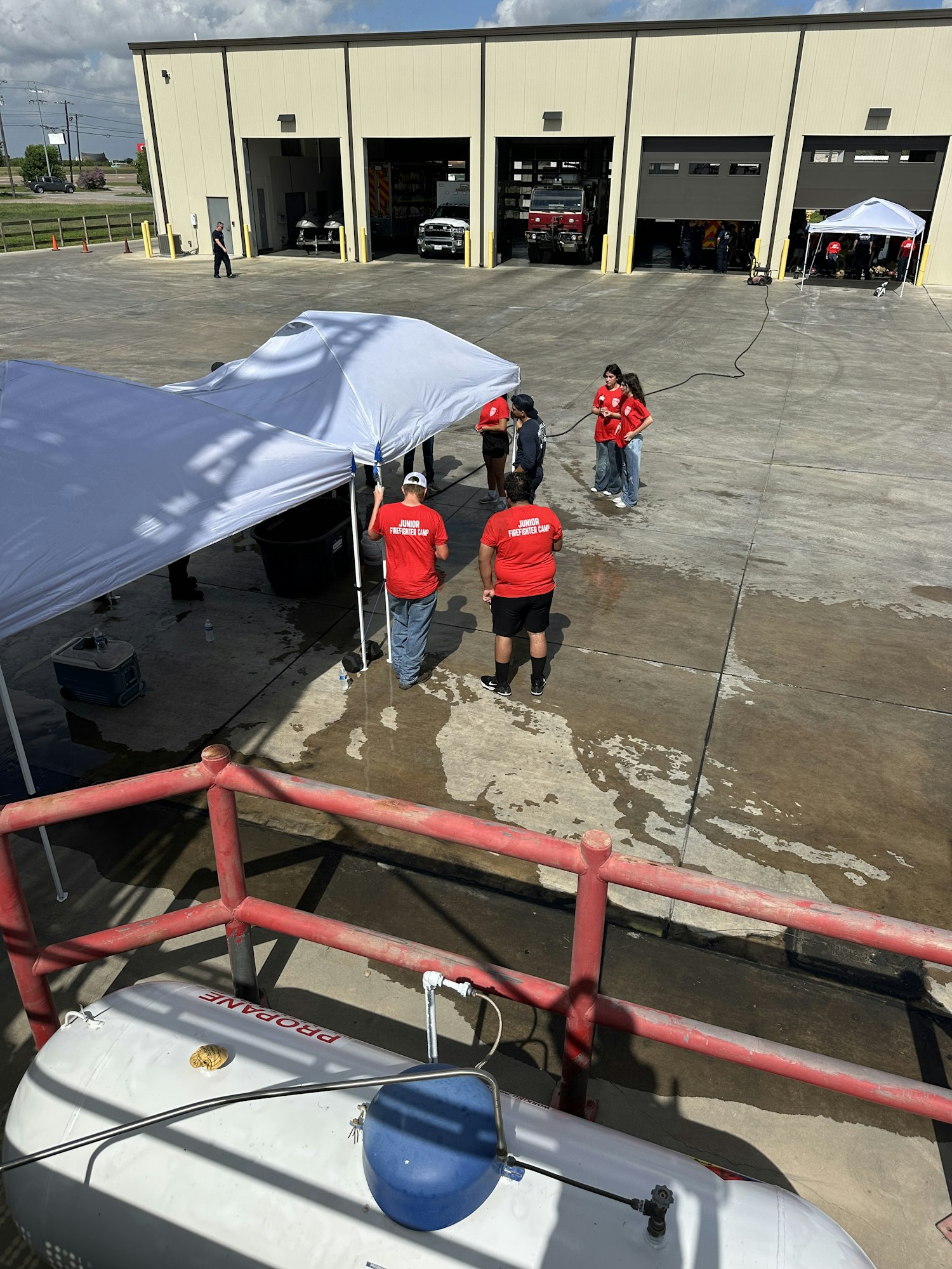 People in red shirts setting up equipment under a canopy near a building with vehicles inside open bays.
