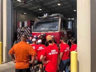 A group of people in red shirts labeled "Junior Firefighter Camp" are gathered around a fire truck inside a station.
