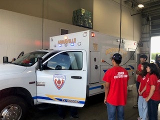 An ambulance parked inside a garage with people in red shirts labeled "Junior Paramedic Camp" standing nearby.
