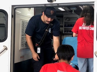 A person in a navy uniform and two people in red shirts inside an ambulance, likely during a junior firefighter camp event.