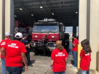 A group in red "Junior Firefighter Camp" shirts standing near a fire truck inside a garage.