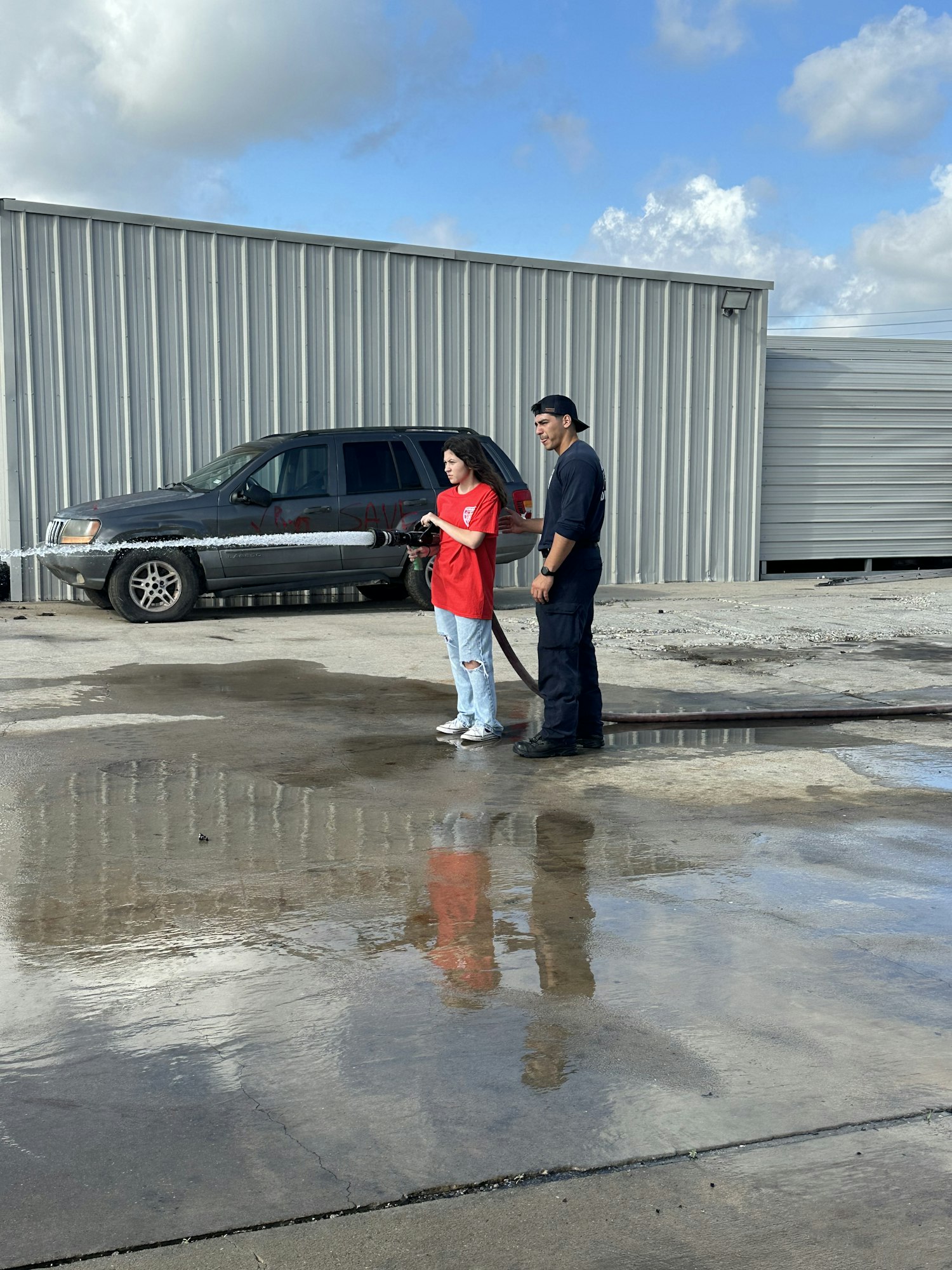 Two people are standing outside by a building, one is holding a hose spraying water.