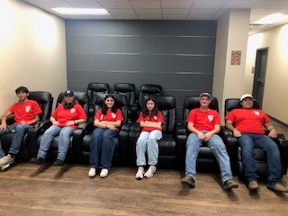 Six people wearing red shirts sitting on black reclining chairs in a room with wooden floor.