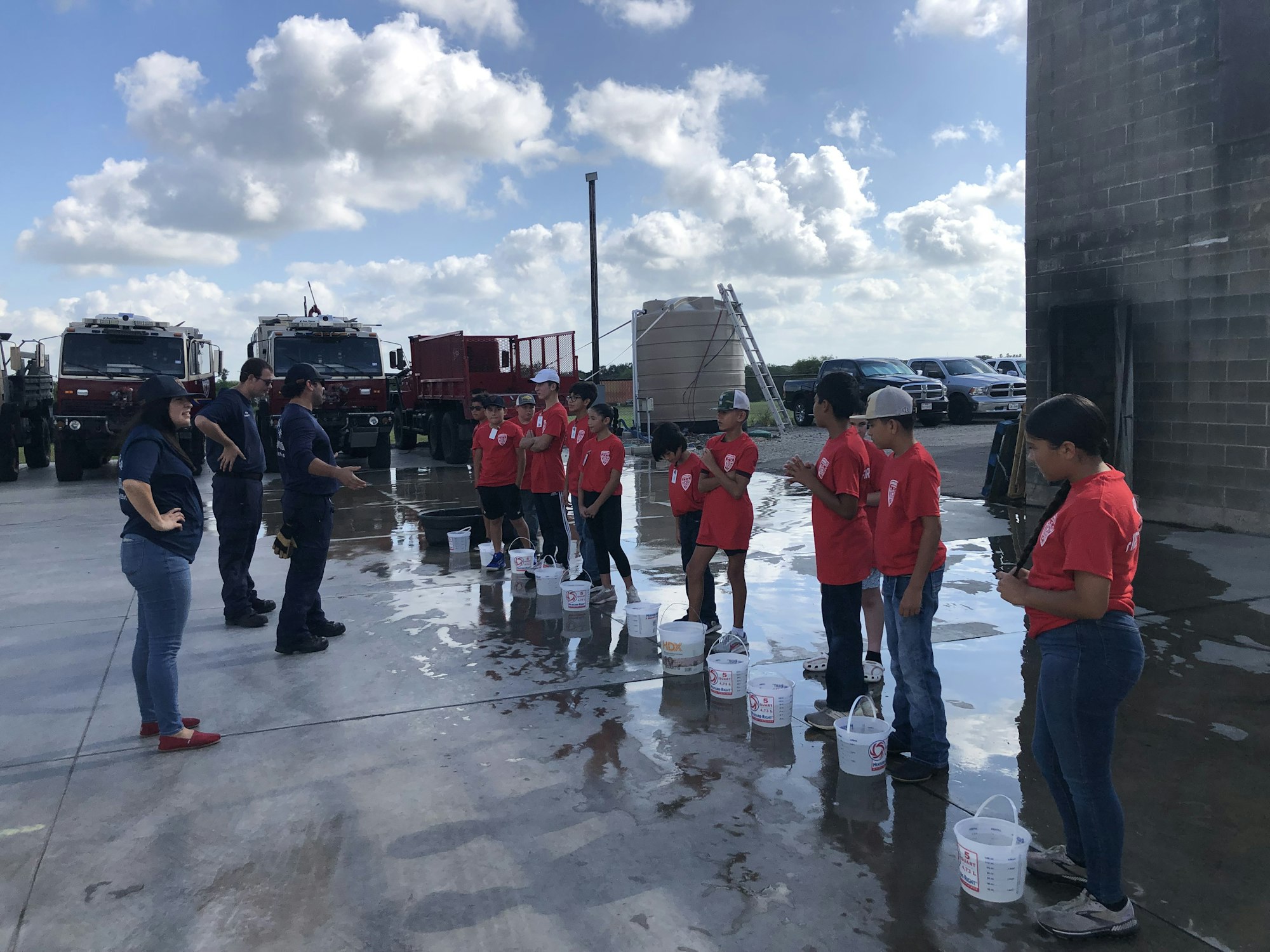 A group of people in red shirts stands in line with buckets, possibly participating in a training or activity, near large vehicles.