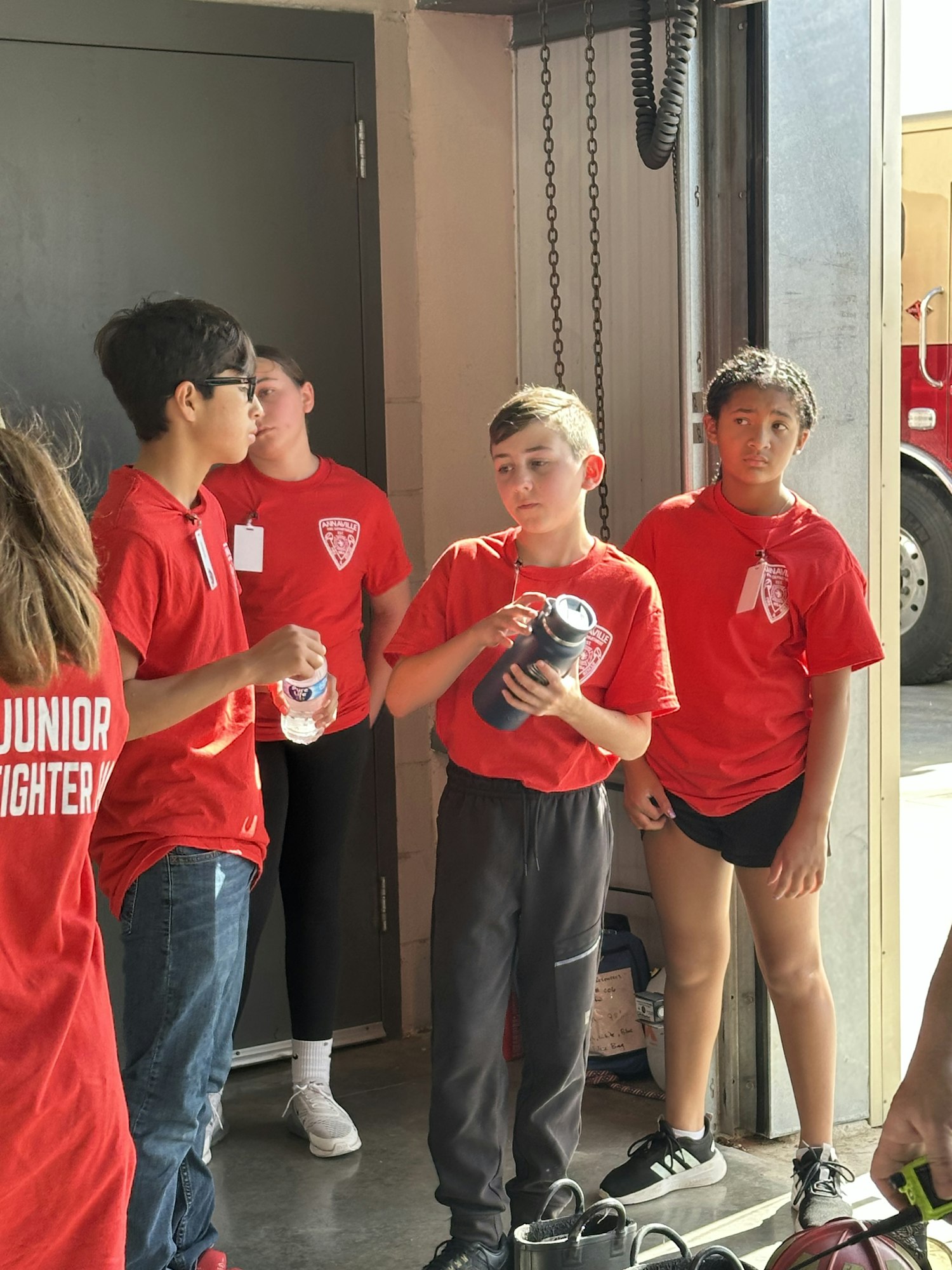 A group of kids wearing red "Junior Firefighter" shirts, standing indoors, holding water bottles.
