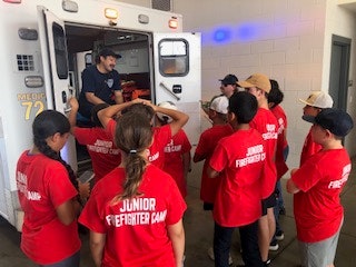 A group of kids in red "Junior Firefighter Camp" shirts listen to a person speaking from an ambulance.