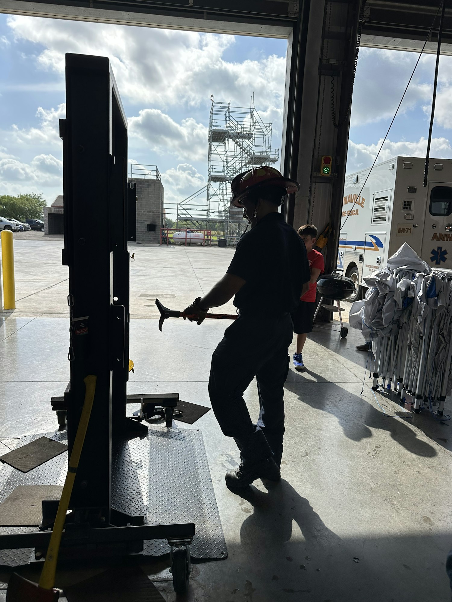 A firefighter holding a tool, standing near an equipment rack, with an ambulance and scaffolding visible outside.