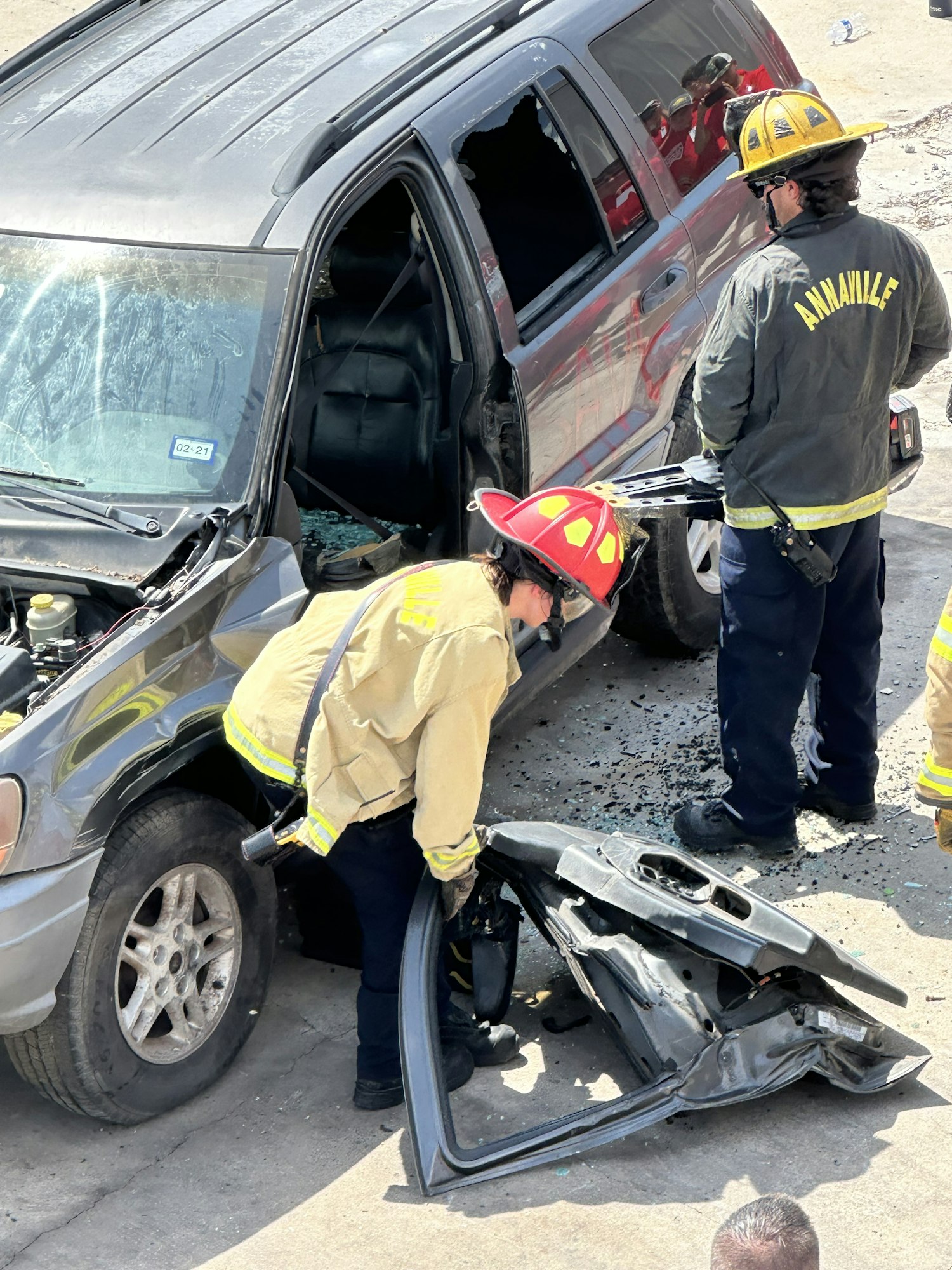 Firefighters work on a damaged vehicle at an accident scene; one holds a detached car part.