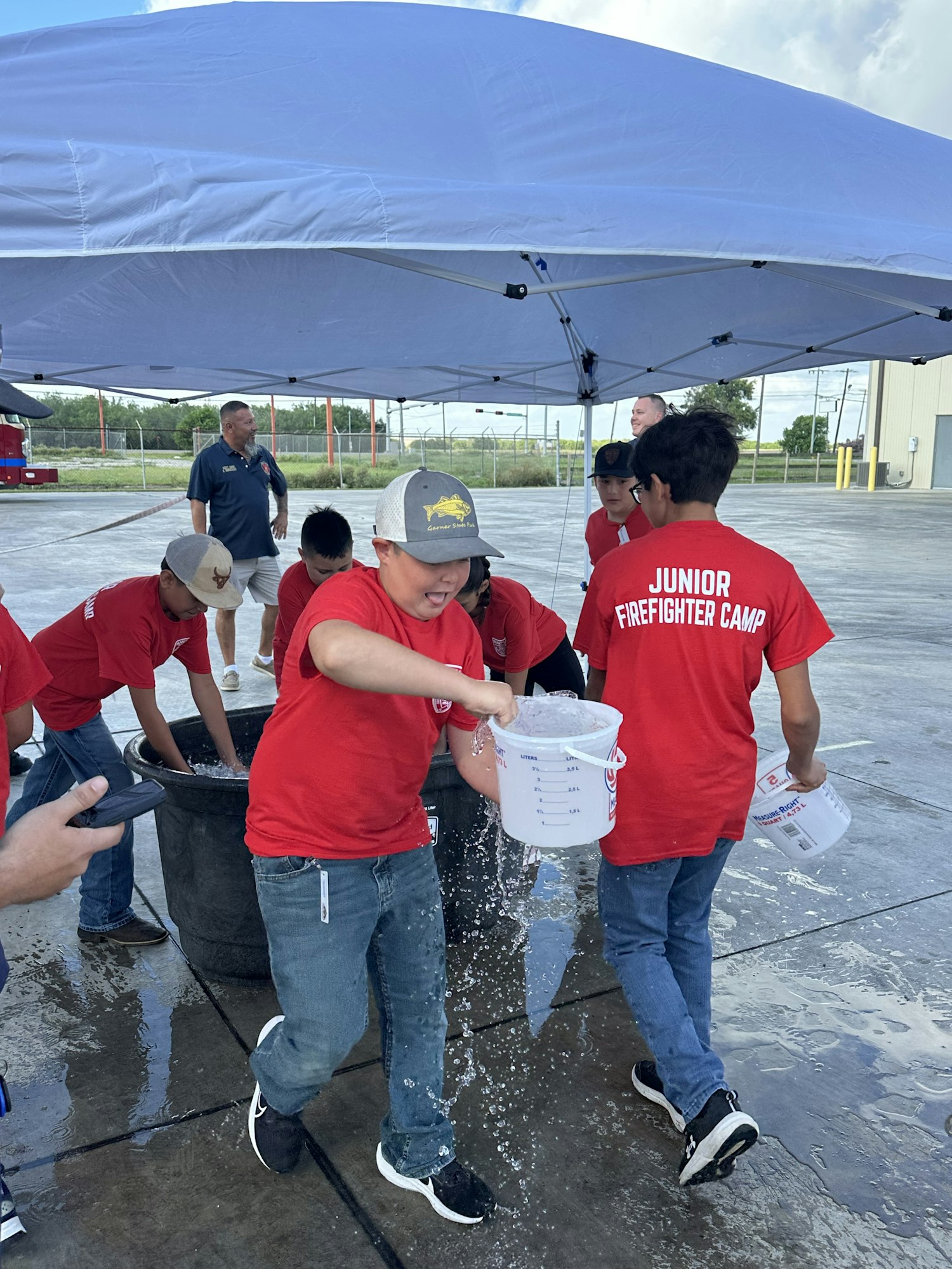 People in red shirts at an event, pouring water into a large container under a canopy. Shirts read "Junior Firefighter Camp."