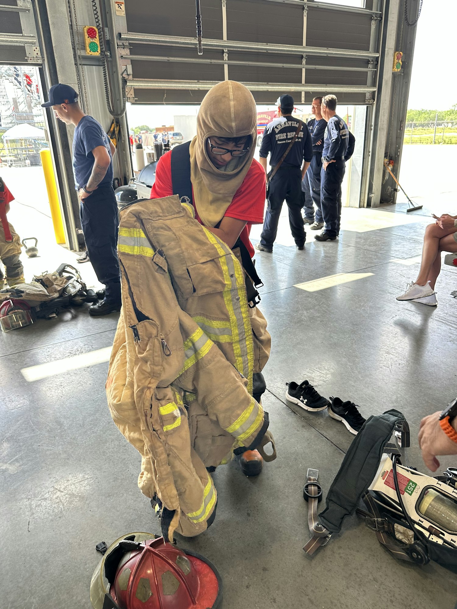 A person dons firefighting gear in a garage while others stand nearby.