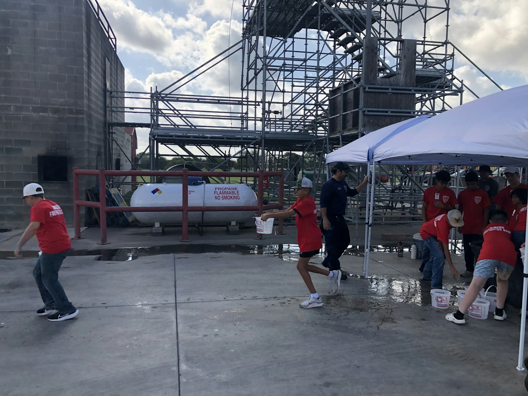 People in red shirts are participating in an outdoor activity involving water buckets, near scaffolding and a propane tank.