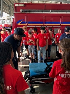 Children in red shirts are gathered around a fire truck at what looks like a fire station event or camp.