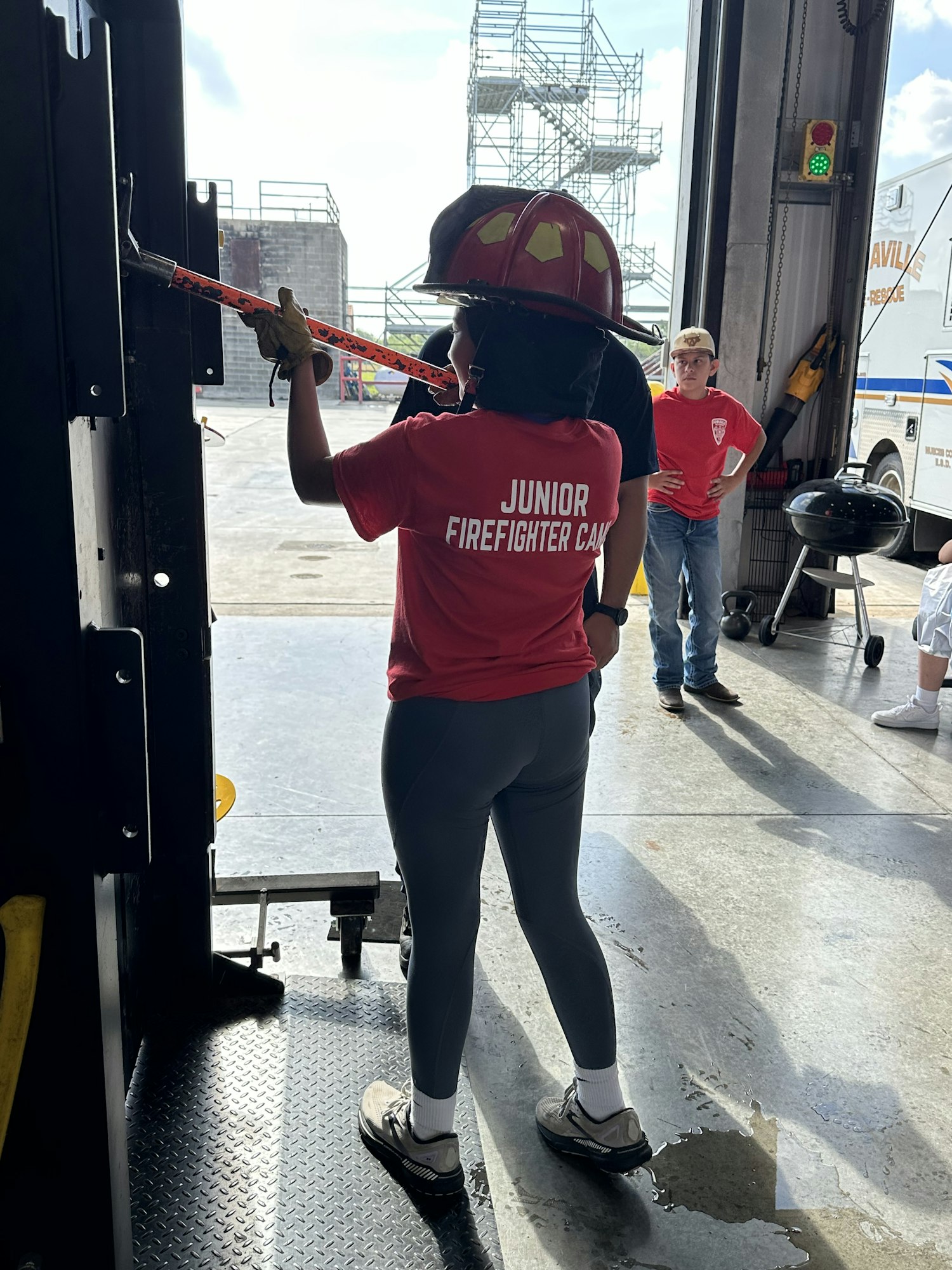 Person in a "Junior Firefighter Camp" shirt using a tool in a garage area, with others present.