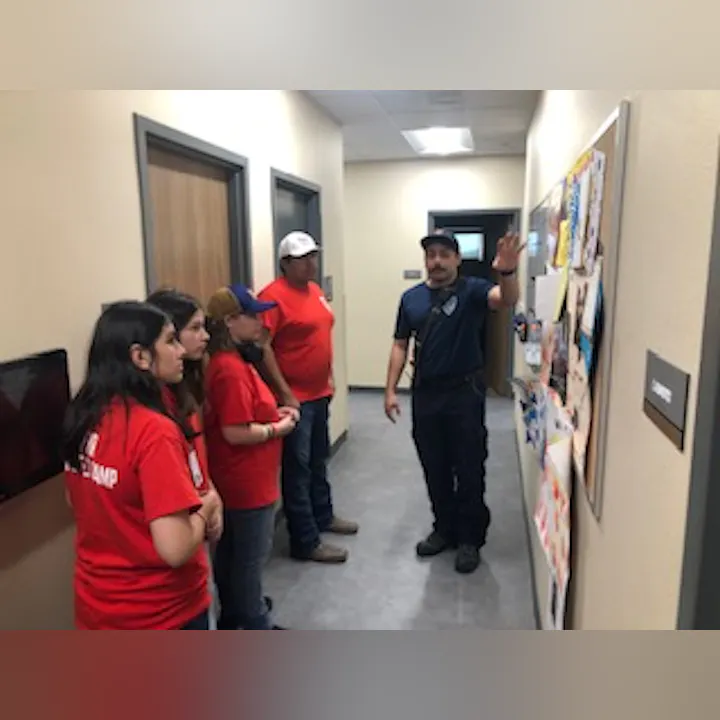 A person in uniform is explaining something on a bulletin board to a group of four people wearing red shirts in a hallway.