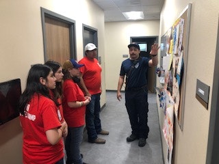 A person in uniform is explaining something on a bulletin board to a group of four people wearing red shirts in a hallway.