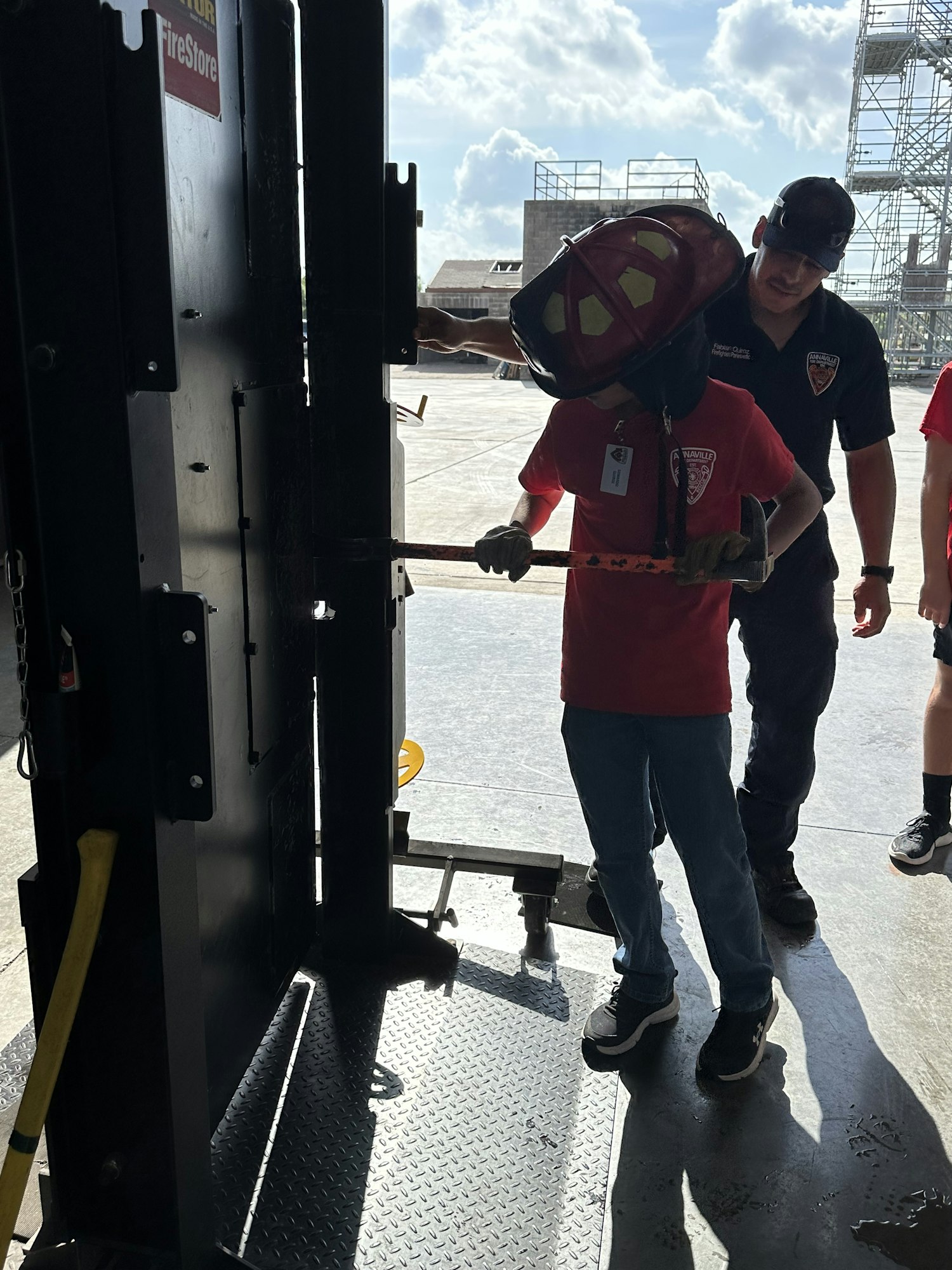 A firefighter instructing a person holding an axe, standing in front of a door breaching simulator.